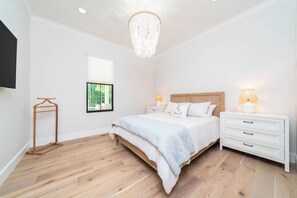 A bedroom with a wooden bed, white nightstands, a hanging light fixture, and a window with a view of greenery. - Luxury-private pool-across from beach (South Haven)
