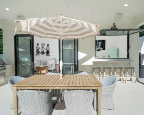 Um pátio externo com mesa de madeira e cadeiras de vime, um guarda-sol listrado e uma porta de vidro que dá para uma cozinha.