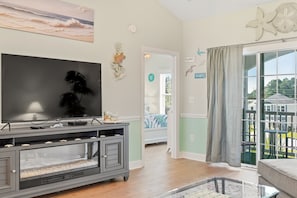 A living room with a flat-screen TV, a grey entertainment unit, and a view of the outdoors through a glass door. - Footprints on the Fairway , 2BR Beach & Golf with Pool (Little River)