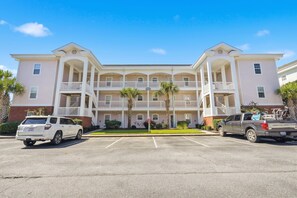 A multi-story hotel with a central courtyard, parking spaces, and a clear blue sky. - Footprints on the Fairway , 2BR Beach & Golf with Pool (Little River)