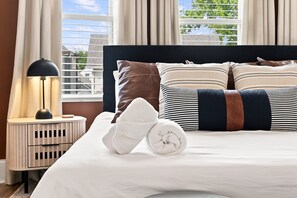 A neatly made bed with a white towel and rolled towel, a black lamp on a bedside table, and a window with blinds. - 5 Min to Lucas Oil - Hot Tub - Arcade - Outdoor Movie - Propane Firepit (Indianapolis)
