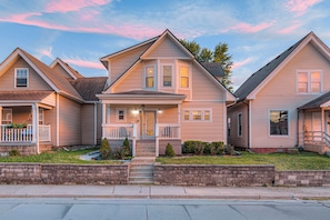 A two-story residential house with a front porch, a small front yard, and a brick retaining wall. - 5 Min to Lucas Oil - Hot Tub - Arcade - Outdoor Movie - Propane Firepit (Indianapolis)