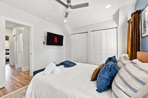 Un dormitorio con cama, ventilador de techo, televisor montado en la pared y una puerta con perchero.