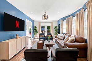 A modern living room with a flat-screen TV mounted on the wall, a brown leather sofa, a wooden coffee table, and a blue accent wall. - 5 Min to Lucas Oil - Hot Tub - Arcade - Outdoor Movie - Propane Firepit (Indianapolis)