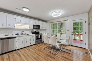 A modern kitchen with white cabinetry, a stainless steel dishwasher, a built-in oven, and a microwave. There is a dining area with a glass table and white chairs. - The Brick House on Elliott (Clarksville)