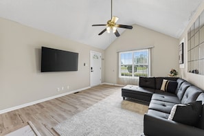 A spacious living room with a flat-screen TV, a sectional sofa, a ceiling fan, and a large window with blinds. - The Brick House on Elliott (Clarksville)