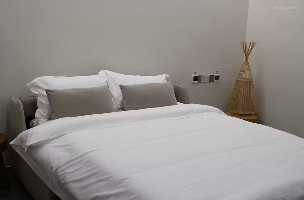 A neatly made bed with white bedding, a grey headboard, and a bedside lamp with a woven base.