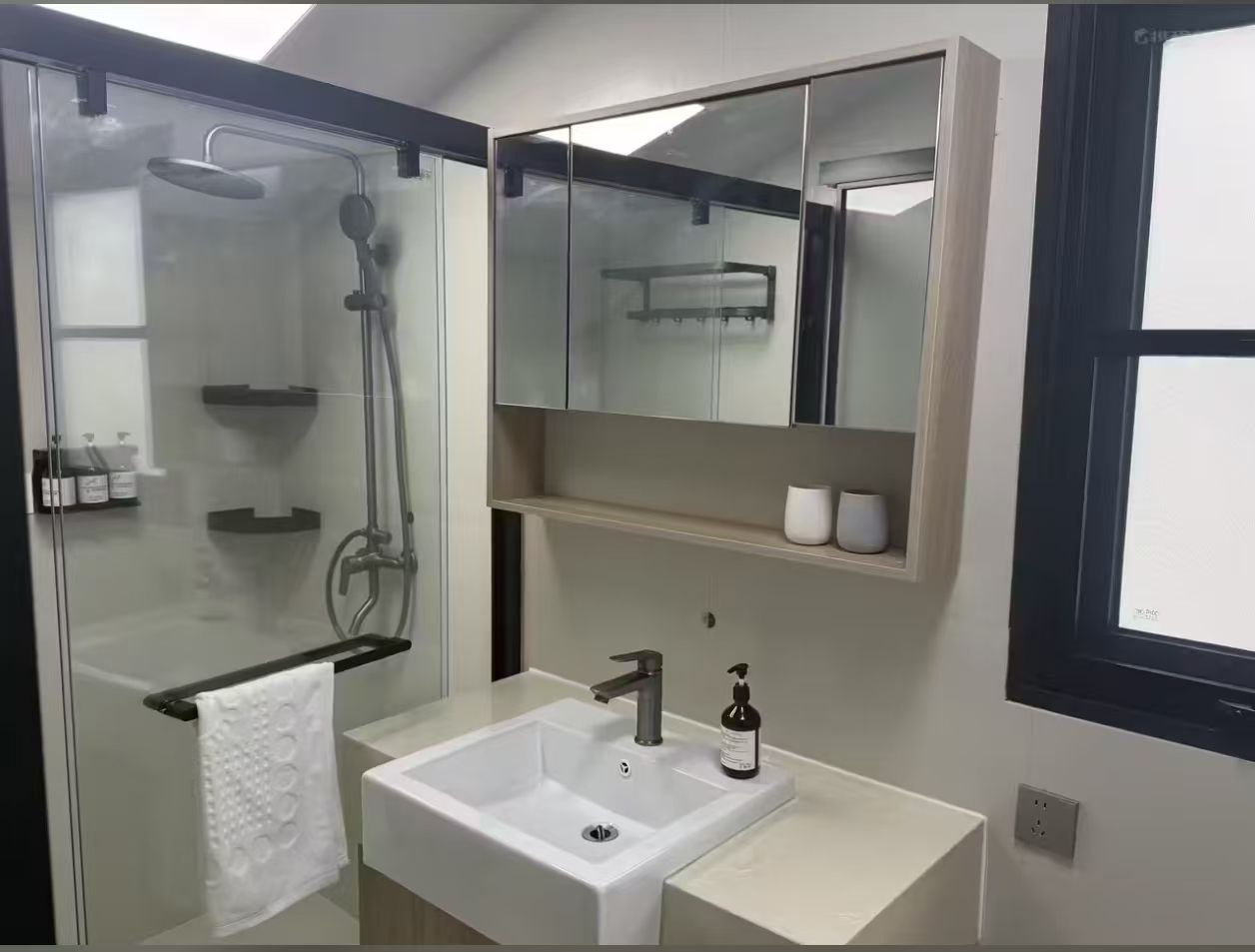 Un baño moderno con cabina de ducha de vidrio, un lavabo blanco con espejo y un estante con artículos de aseo.
