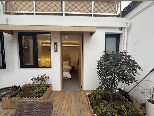 A two-story building with a wooden deck, a bed visible through an open door, and a potted plant in the foreground.