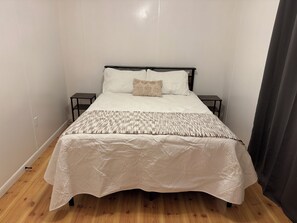 Una cama individual con colcha acolchada y dos almohadas, con mesitas de noche a cada lado, en una habitación con paredes lisas y suelo de madera.