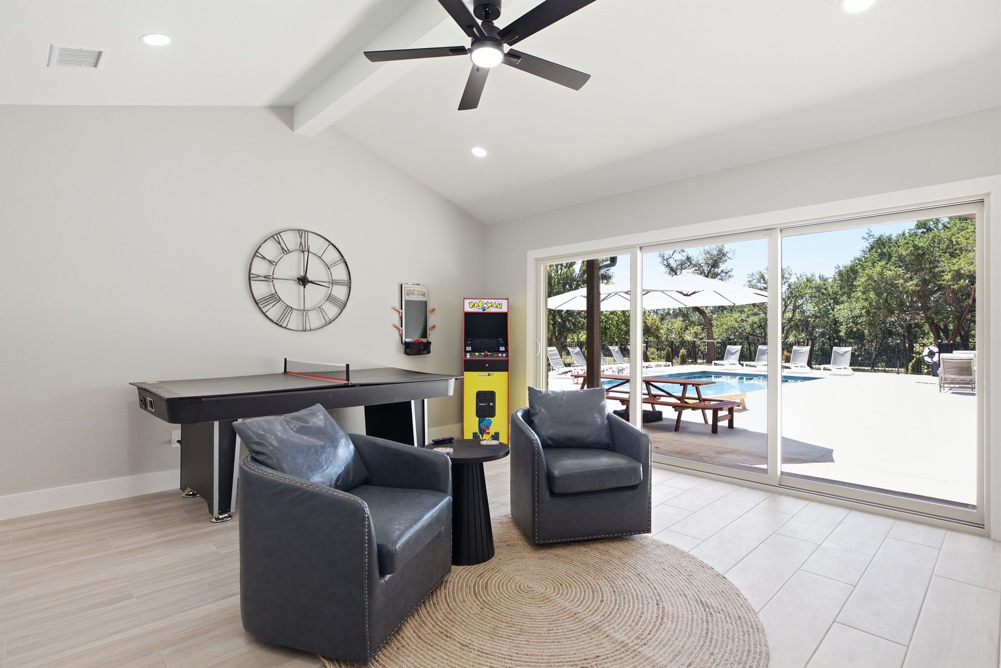 Un salon moderne avec une table de billard, deux fauteuils, un ventilateur de plafond et une grande fenêtre donnant sur une piscine.