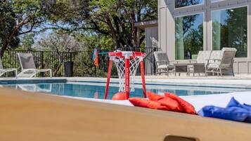A swimming pool with a basketball hoop, lounge chairs, and a building with large windows in the background.