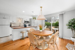 A modern kitchen with white cabinets, a central island, and a dining area with wicker chairs and a wooden table. - Ripple Effect - near the beach and Main Street (Hyannis)