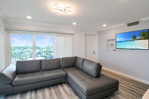 Ein modernes Wohnzimmer mit einer grauen Eckcouch, einem großen Fernseher an der Wand und einem Balkon mit Blick auf Grünflächen.