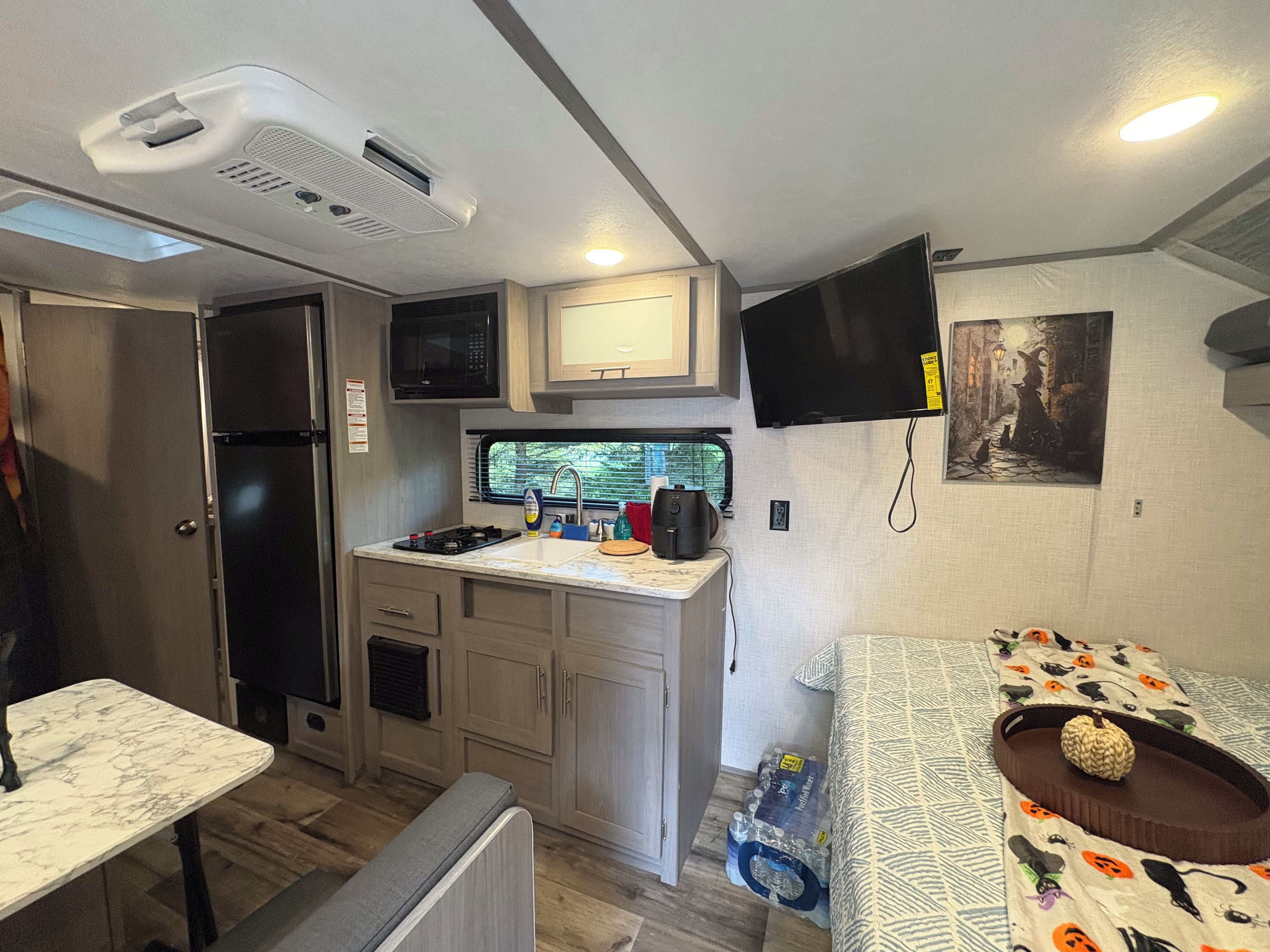 A compact RV kitchen with a microwave, sink, and stove, a television mounted above the counter, and a bed with Halloween-themed bedding.