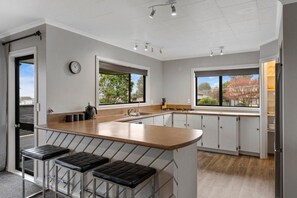 A modern kitchen with a central island, bar stools, a clock, and large windows offering views of the outside. - Taupō Group Getaway | 6 Bedrooms, 3 Bathrooms (Taupō)