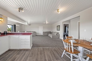 Eine moderne Küche mit weißen Schränken und einer roten Fliesenfassade, ein Essbereich mit Holztischen und -stühlen sowie ein Wohnzimmer mit grauem Sofa und großem Fenster.