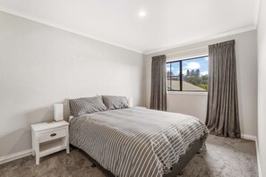 A bedroom with a bed, a nightstand, a window with curtains, and a view of the outdoors. - Taupō Group Getaway | 6 Bedrooms, 3 Bathrooms (Taupō)