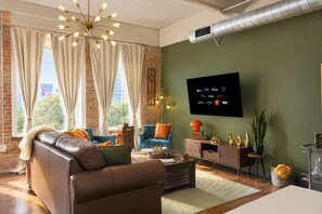 A modern living room with a leather sofa, a blue armchair, and a coffee table. A flat-screen TV mounted on the wall displays various streaming services. - The Bentley Collection - The Retro Retreat (Dallas)