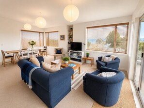 A modern living room with a sofa set, a coffee table, and a TV. There are bookshelves and a dining area visible. - Modern Family Bach | Walk to the Lake | Sleeps 8 (Taupō)