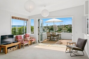A modern living room with a flat-screen TV, wicker chairs, a dining table, and a large umbrella. - Modern Family Bach | Walk to the Lake | Sleeps 8 (Taupō)