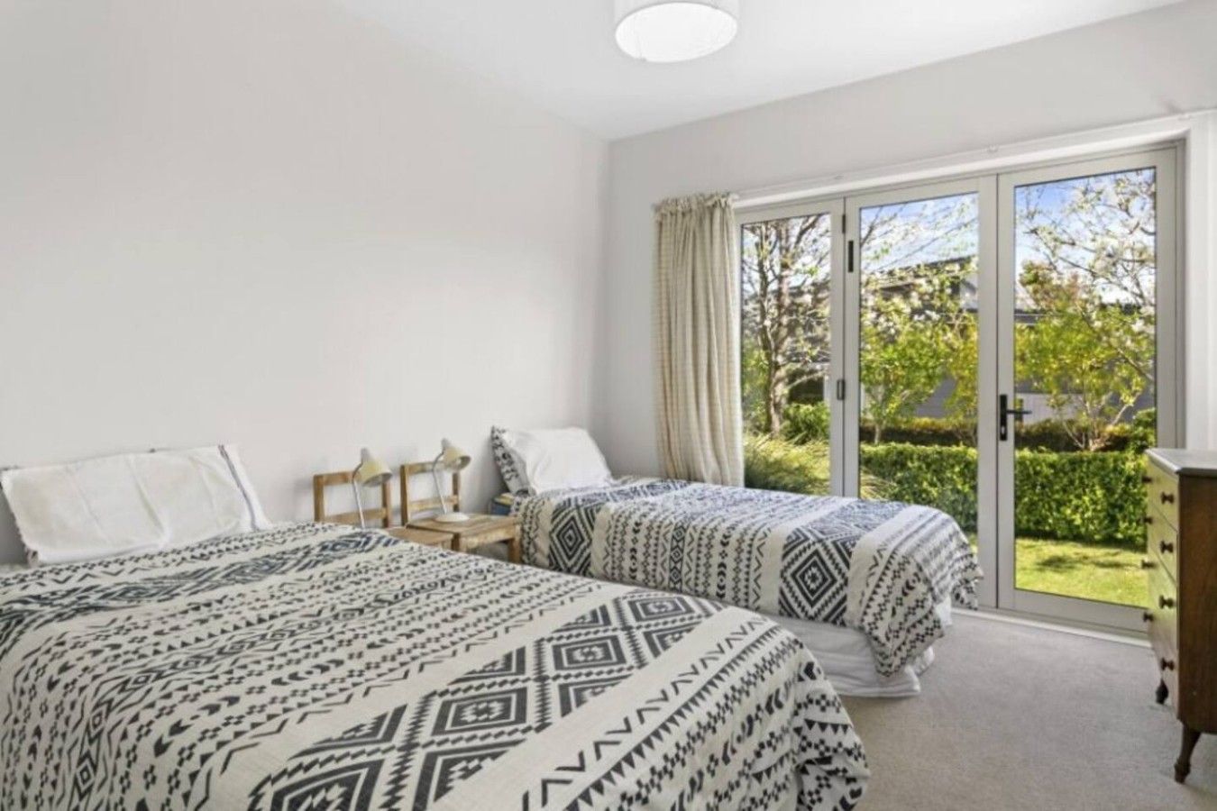 A bedroom with two beds, a nightstand, a dresser, and a sliding glass door leading to a garden.