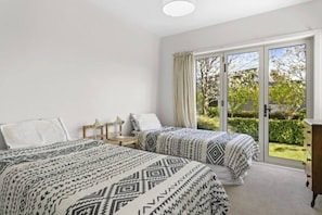 A bedroom with two beds, a nightstand, a dresser, and a sliding glass door leading to a garden. - Modern Family Bach | Walk to the Lake | Sleeps 8 (Taupō)