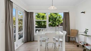 A room with a dining table and chairs, large windows with curtains, a hanging light fixture, and a small plant on a cart.