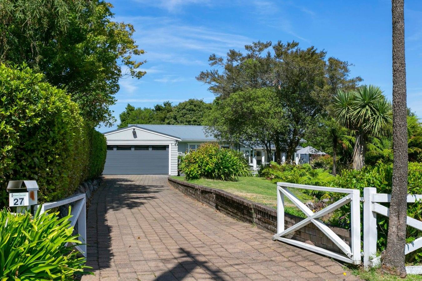 A residential property with a paved driveway leading to a garage, surrounded by lush greenery and a white fence.