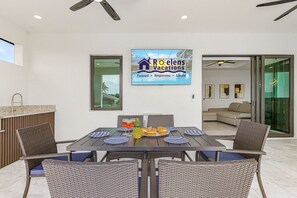 Ein Essbereich mit einem Tisch für vier Personen, einem Schild für Roelens Vacations sowie Blick auf einen Pool und Palmen.