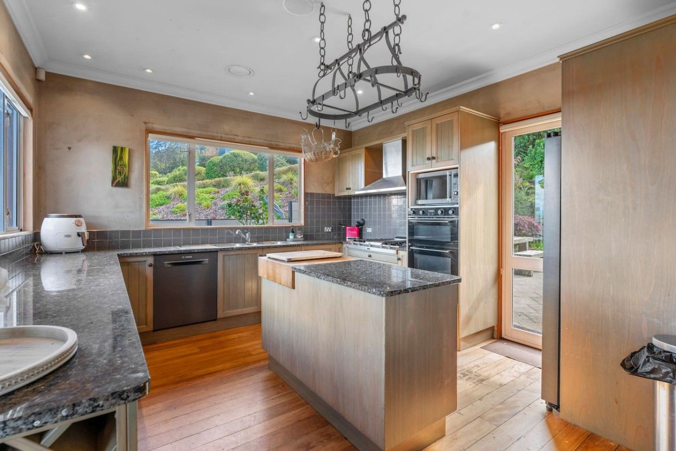 A modern kitchen with a central island, stainless steel appliances, and a view of a garden through large windows.