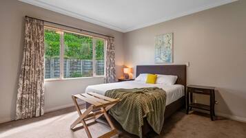 Ein Schlafzimmer mit einem Bett, einem Nachttisch, einer Lampe, einem Fenster mit Vorhängen, einem Holzstuhl und einem kleinen Tisch.