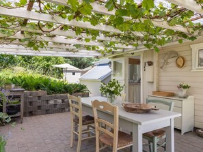 Eine überdachte Terrasse mit Tisch und Stühlen, eine weiße Wand mit einem Fenster und ein durch eine offene Tür einsehbarer Küchenbereich.