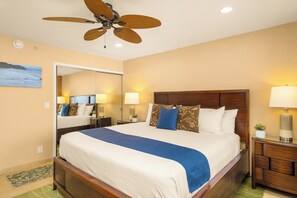 A hotel room with a bed, a ceiling fan, a nightstand, a lamp, a mirror, and a view of the ocean through the window. - Beautiful 1BR Oceanfront Retreat in Kaanapali (Lahaina)