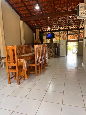 A dining area with a long wooden table and chairs, a tiled floor, a television, and a kitchen area with a refrigerator. - House in a Gated Community - Caldas Novas, Goiás (Caldas Novas)