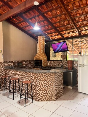 A kitchen with a checkered tile backsplash, a built-in oven, a bar area with stools, and a television mounted on the wall. - House in a Gated Community - Caldas Novas, Goiás (Caldas Novas)