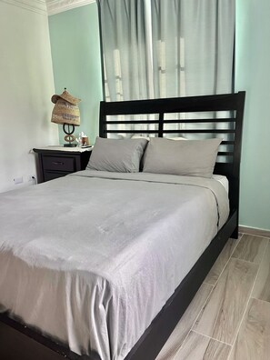 Ein ordentlich bezogenes Bett mit Kopfteil, ein Nachttisch mit Lampe und eine Steckdose an der Wand.