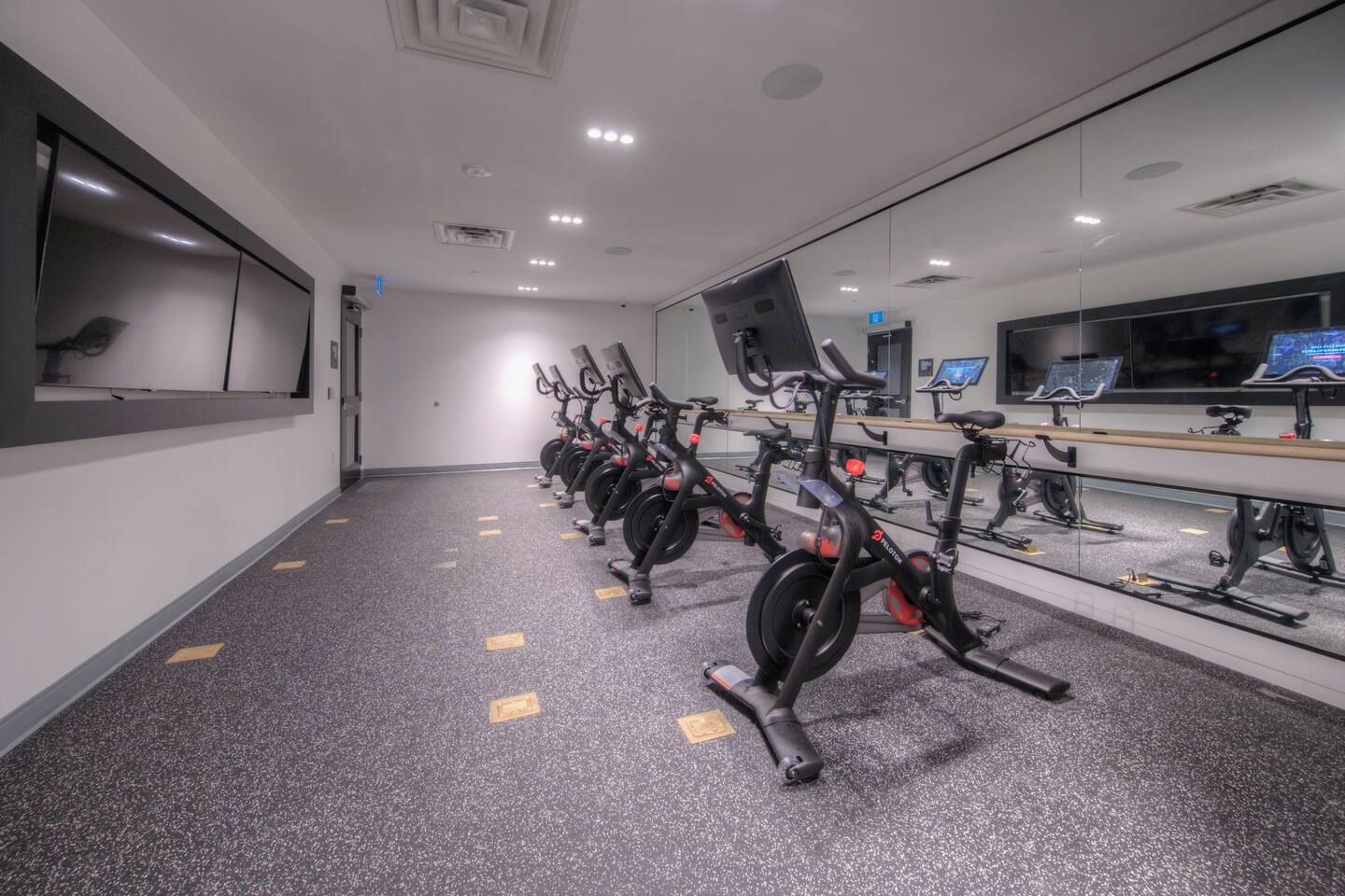 Uma academia com bicicletas ergométricas dispostas em linha, um espelho grande e uma televisão presa à parede.