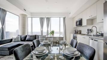 A modern living room with a glass dining table set for four, a sofa, and a kitchen area with white cabinets and stainless steel appliances.