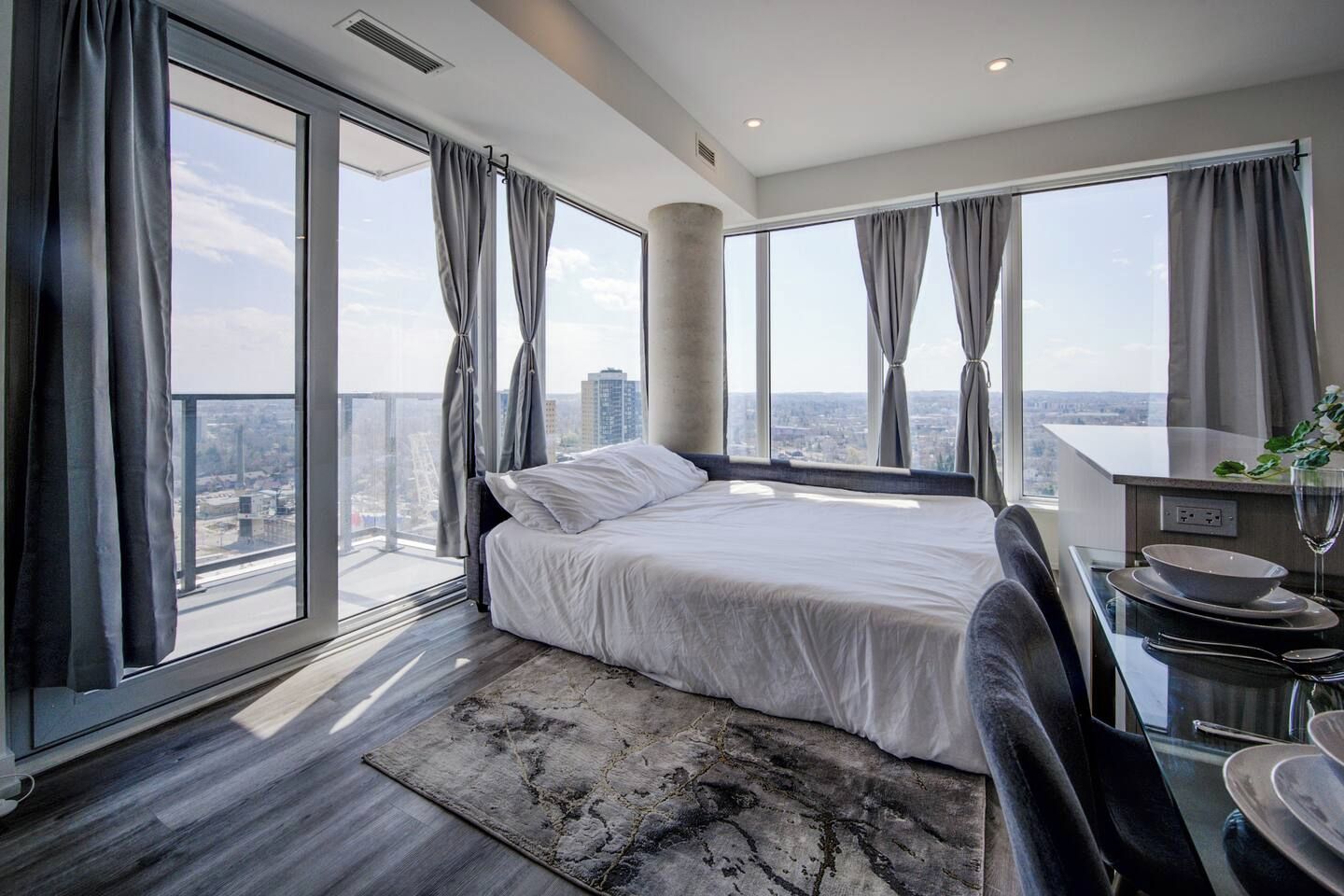 A modern bedroom with a large bed, a dining area with a table and chairs, and a view of the city through large windows.