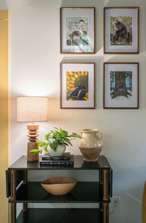 Eine Wand mit vier gerahmten Bildern, einer Lampe, einem Blumentopf und einem kleinen Tisch mit Büchern und einer Keramikkanne.