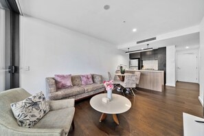 Un salon moderne comprenant un canapé, un fauteuil et une table basse, un coin cuisine avec un bar, et une porte donnant sur une autre pièce.