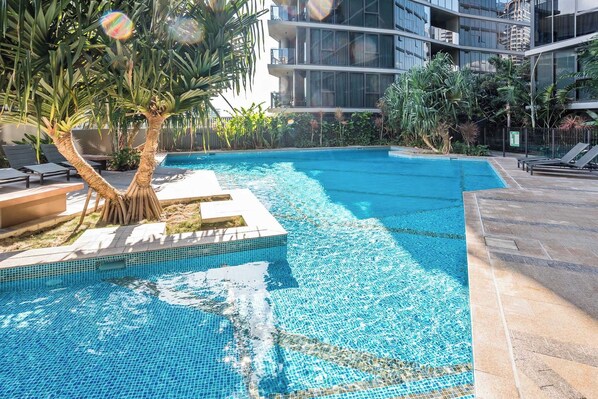 Uma piscina com borda azulejada, cercada por cadeiras de descanso e uma palmeira.