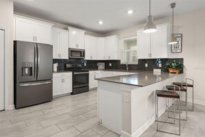 A modern kitchen with white cabinetry, stainless steel appliances, a central island with bar stools, and pendant lighting. - Private Pool & Spa | Six-Bedroom ChampionsGate Home (Kissimmee)