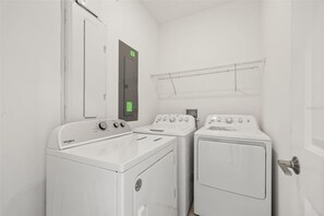 A laundry room with two washers and a dryer, a metal rack, and a door with a green sign. - Private Pool & Spa | Six-Bedroom ChampionsGate Home (Kissimmee)