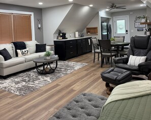 A modern living room with a sofa, coffee table, and ottoman, a dining area with chairs and a table, and a kitchen with cabinets and appliances.