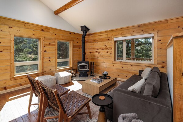 A cozy cabin interior with a fireplace, wooden walls, a sofa, a coffee table, and large windows.