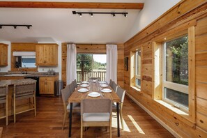 A wooden cabin interior with a dining table set for six, kitchen area, and large windows.