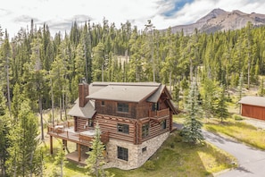 A log cabin with a stone foundation, a deck, and a mountain backdrop. - New! Ski-in/Ski-out, Private Hot Tub, BBQ, Lone Peak! (Big Sky)