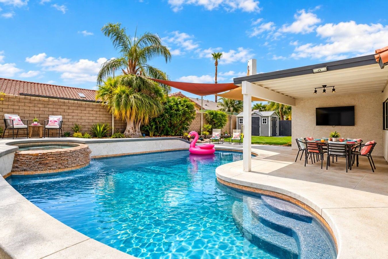 A swimming pool with a stone-lined edge, a pink flamingo float, and a covered patio area with outdoor furniture.